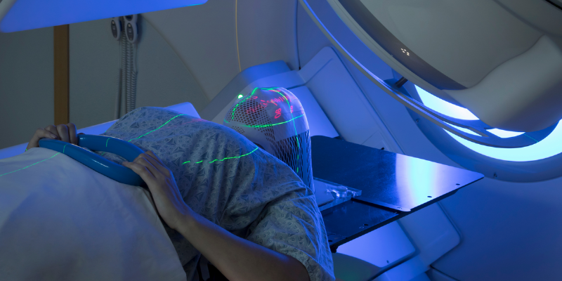 A woman lies on a hospital bed underneath a radiotherapy machine, wearing a blue hospital gown