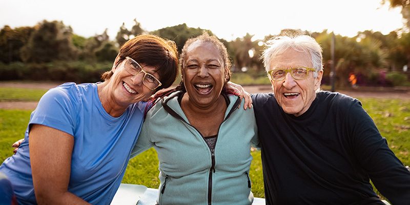 Three mature people sat smiling, arm in arm, in a park.