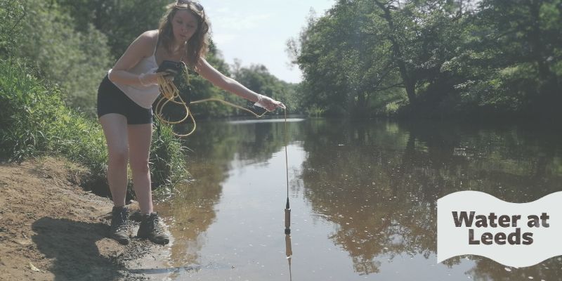 Maddy Wright holding a testing probe in the River Nidd. The wave-shaped logo in the corner reads Water at Leeds.