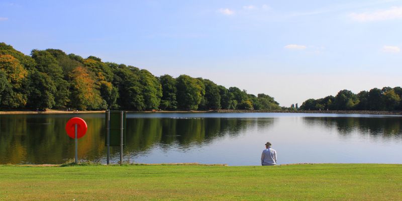 A figure stands by the lake in Roundhay Park in Leeds. The lake is reflecting the bright blue sky.