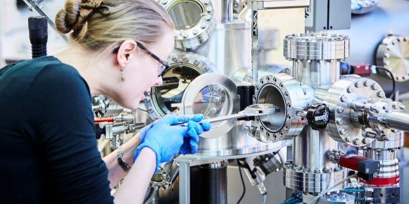A scientist wearing safety glasses and blue gloves carefully operates a complex laboratory vacuum chamber and scientific instrumentation during a research experiment.
