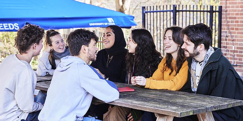 Seven students smiling and laughing, sat around a table outside the Leeds University Union.