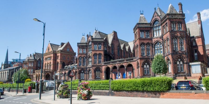 Historic red-brick hospital building at Leeds General Infirmary, featuring ornate towers, arches and decorative stonework, viewed from a street with landscaped flower displays and parked cars in the foreground.