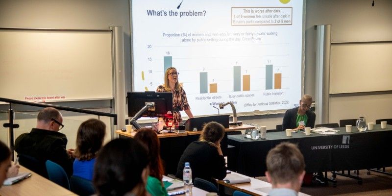 Lecture hall presentation showing a slide titled ‘What’s the problem?’ with a bar chart comparing feelings of safety for women and men in different public spaces. Audience members are seated at desks, and a speaker stands near a podium.