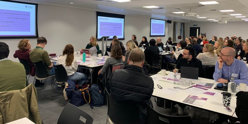 People sit in a white seminar room, taking notes and staring at presentation screens in front of them.