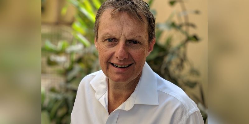 Headshot of man facing the camera. He is wearing a white, open neck shirt. There is foliage behind him