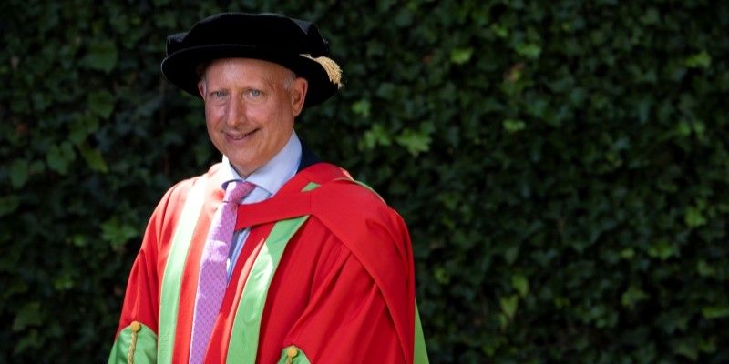 Steve Rosenberg stands smiling at the camera in red and green graduation robes.