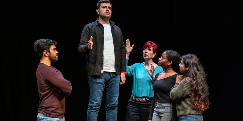 A performance group in a drama space. One person is stood on a raised area and and educator is directing.