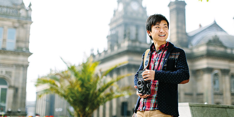 A smiling student with a camera in Leeds city centre. Leeds Town Hall is in the background.