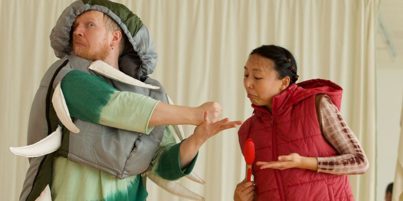 A man wearing a green insect costume on the left is looking away from the woman on the right, who is wearing a red jacket. They play rock, paper, scissors.
