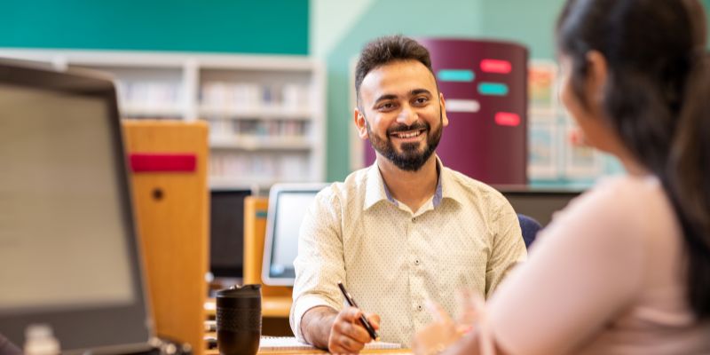 A student chatting to a member of staff, sat at a computer in a study space.