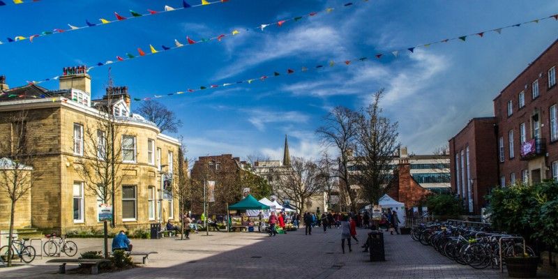 Outside Leeds University Union: an open pedestrian space with buildings on either side and colourful bunting overhead. People are walking around and visiting stalls in gazebos.