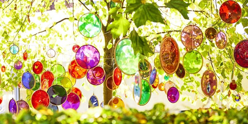 Glass shapes in different vibrant colours hanging in the branches of a tree on a sunny day.