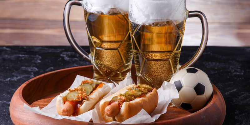 A pair of hotdogs and pints of beer on a wooden board with a mini-football.