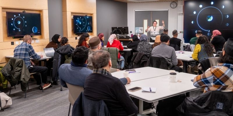 A classroom-style workshop where participants sit at tables listening to a presenter at the front of the room. Large screens on the walls display diagrams of planetary orbits and the Earth–Moon system. Attendees face forward, taking notes.