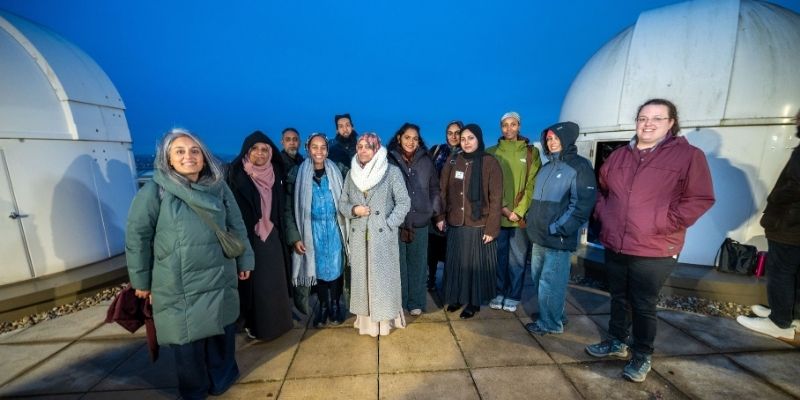 A group of people standing together outdoors on a paved observatory platform at dusk, with two large white domed structures on either side and a deep blue sky in the background. The group is dressed in winter clothing, including coats, scarves, and hats.