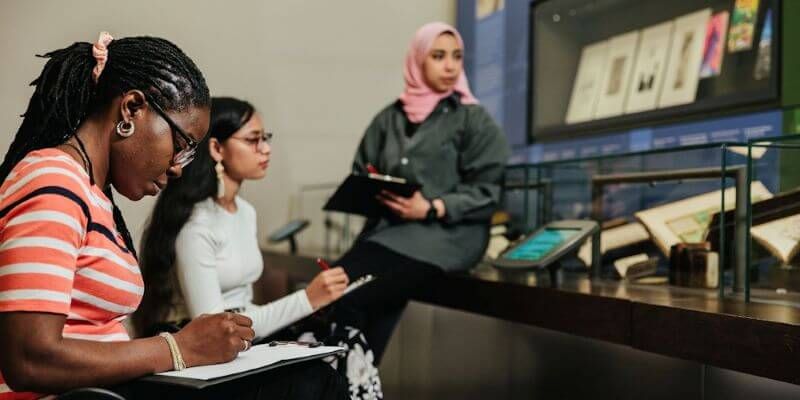 Three women sit writing on notepads, looking at a museum exhibit.