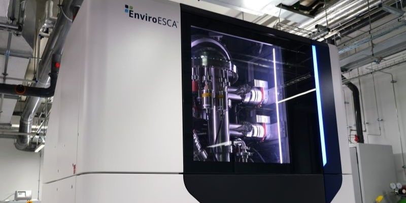 The XPS, a room-height white and black metal structure with glass door panel, featuring the EnviroESCA logo. The instrument’s wiring and mechanics are visible through the glass panel.