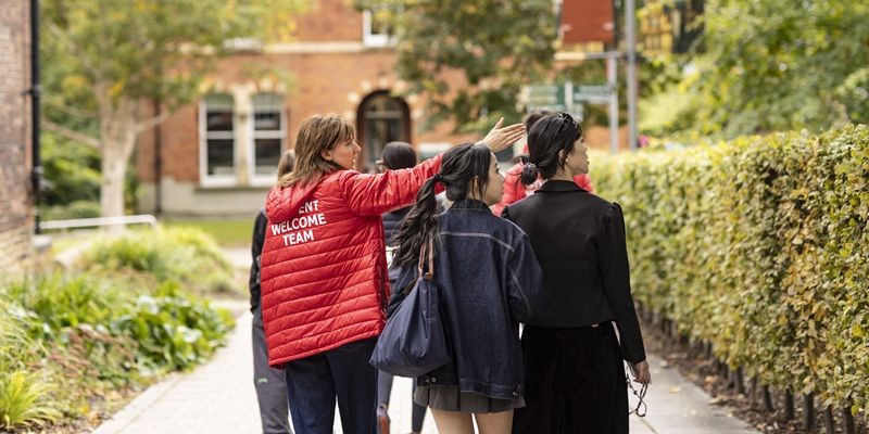 A group of people being shown around campus by a member of the Student Welcome Team. They are looking at something the staff member is pointing to, surrounded by red brick buildings and hedges.