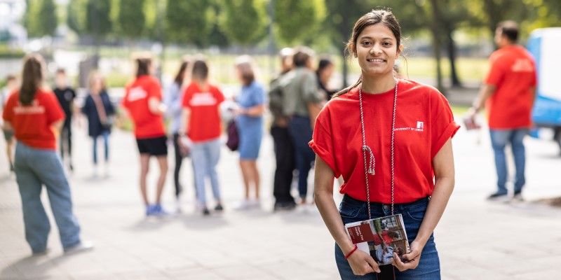 A Leeds student ambassador standing on campus, smiling, holding a brochure, with other student ambassadors and visitors in the background.