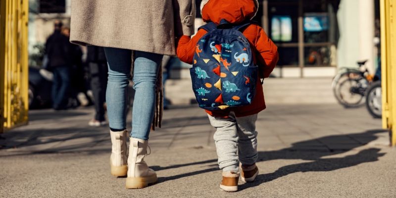 Adult and child walking on a city sidewalk; child wears red coat and dinosaur-print backpack.