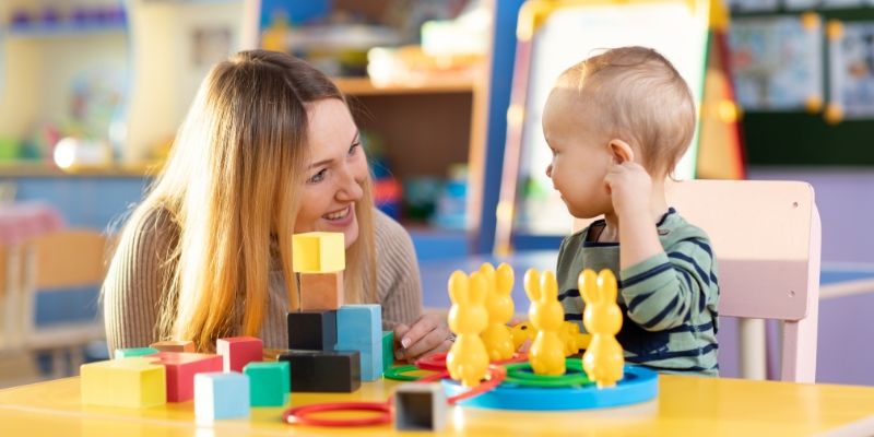 Toddler and childcare worker play with colourful wooden block toys on table