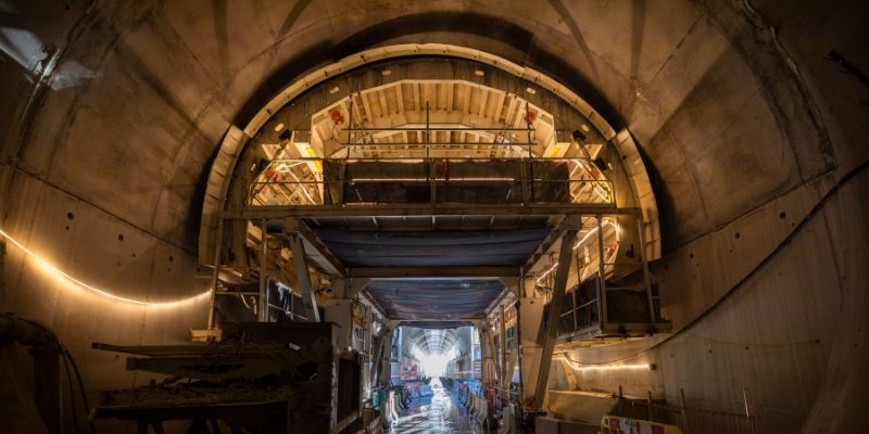Interior view of a large concrete tunnel under construction, showing a multi-level scaffold structure spanning the width of the tunnel, with workers and equipment along a wet walkway and temporary lighting illuminating the curved tunnel walls.