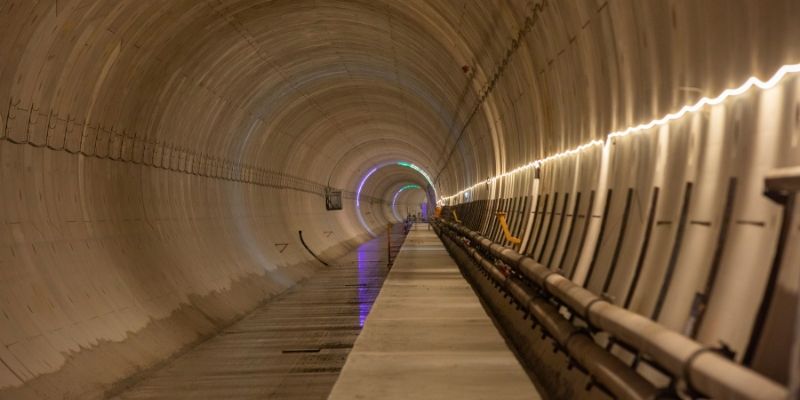 Interior of a large circular tunnel with smooth concrete walls, showing a long walkway and parallel pipes running along the right side, illuminated by continuous strip lighting, with coloured lights visible in the distance.