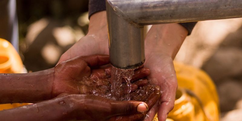 A child holds their hands out under a pipe, from which clean water is flowing. An adult holds their hands out underneath the child’s.