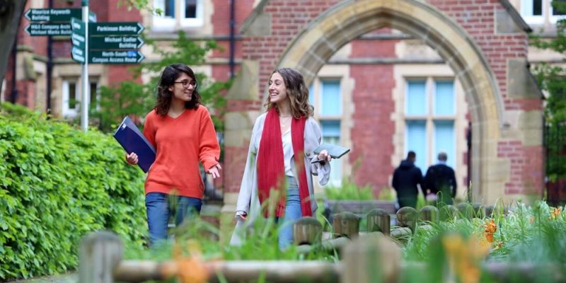 Two students walking and chatting in a green space on campus. They are in front of a decorative red brick archway.