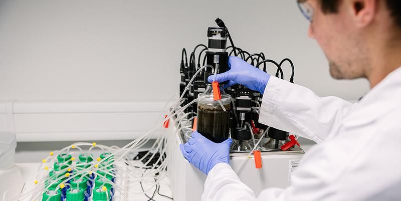 A person in a lab coat and gloves using a machine that is linked to bottles containing liquid