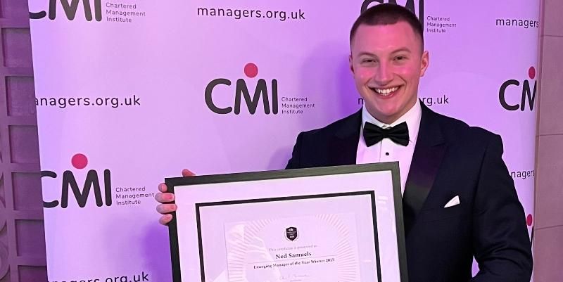 Ned Samuels is stood holding his  framed award in front of a Chartered Management Institute banner.