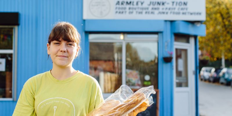 A smiling student volunteer stands holding baguettes in front of Armley Junk-tion cafe.