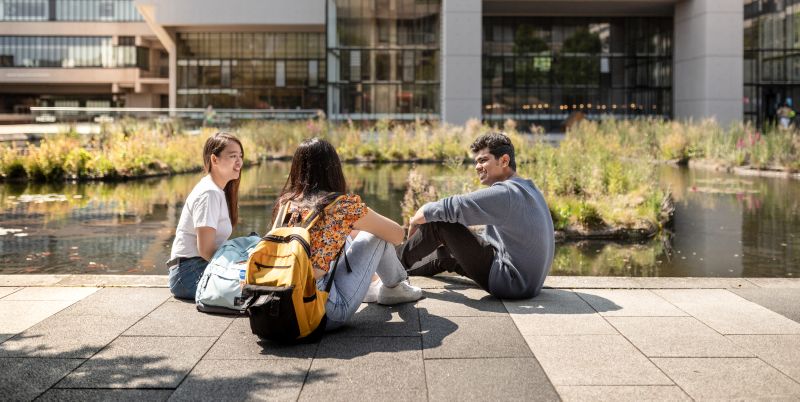 Three students sit next to the Roger Steven's pond in conversation
