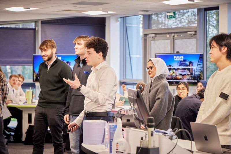 A group of students pitching to a room of people. One student is stood behind the central teaching space computer.