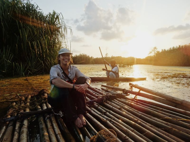 Dr Josie South is sat on a bamboo raft on the water with trees and a sunset behind. 