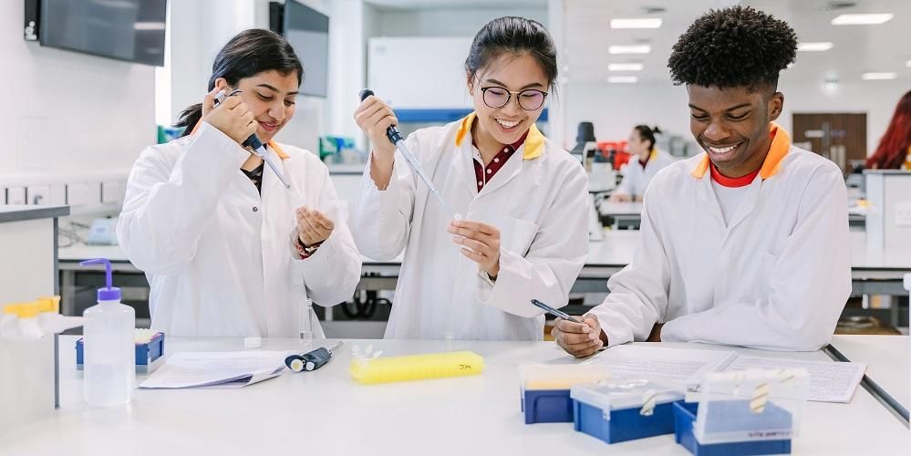 Three students in a laboratory. Two are using pipettes, one is writing.