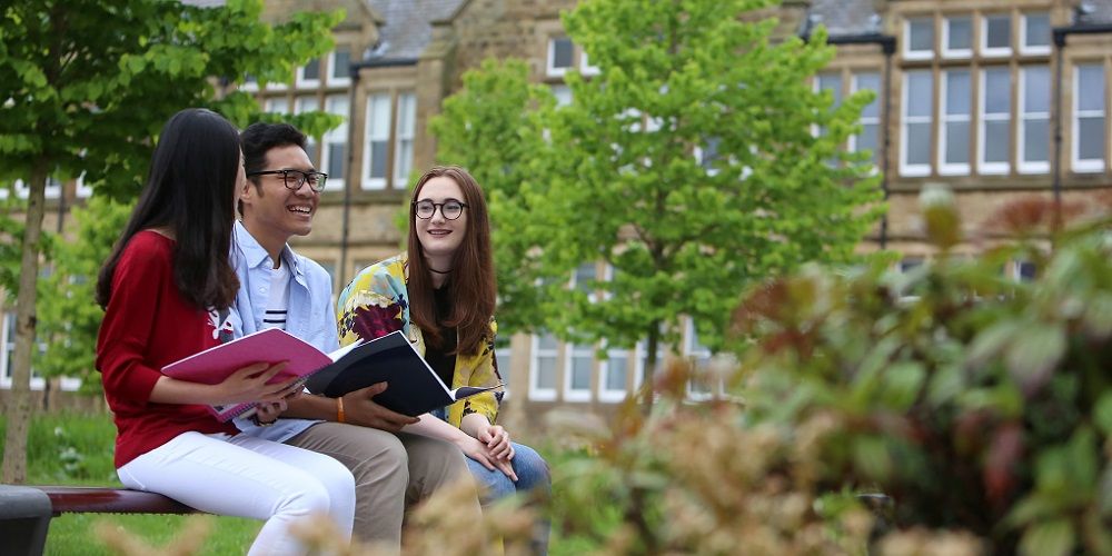 A group of students chatting and smiling, sat on a bench with trees and old buildings in the background.