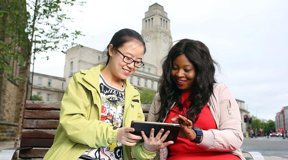 Two students wearing bright clothing sitting on a bench in front of the Parkinson Building reading from a tablet.