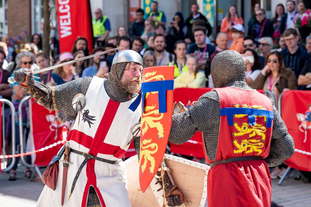 Two knights in chainmail fight with swords in front of a crowd of spectators.