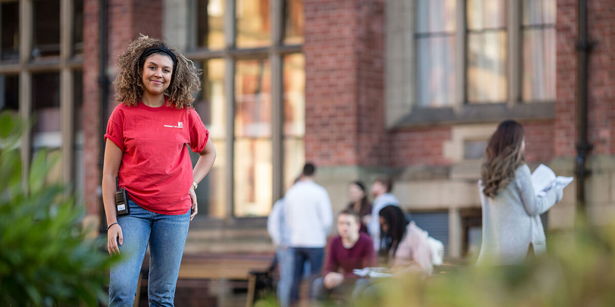 A student open day ambassador is stood on campus smiling. A University building and some open day visitors are in the background