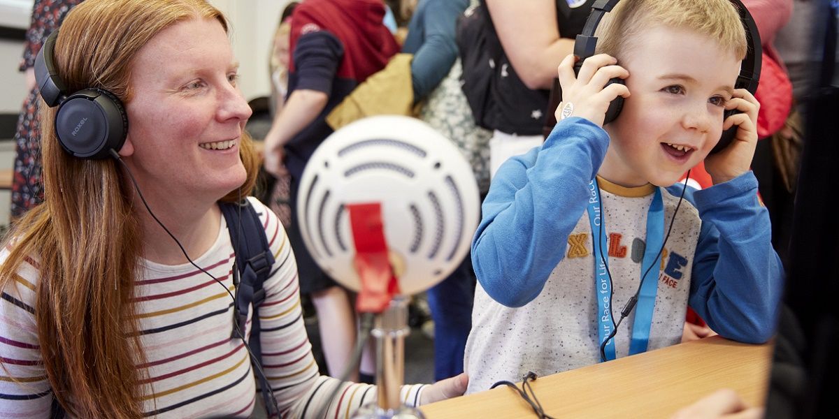 A small child and their parent are sat at a desk listening to something over headphones. The child is holding the headphones over their ears and they both look excited.