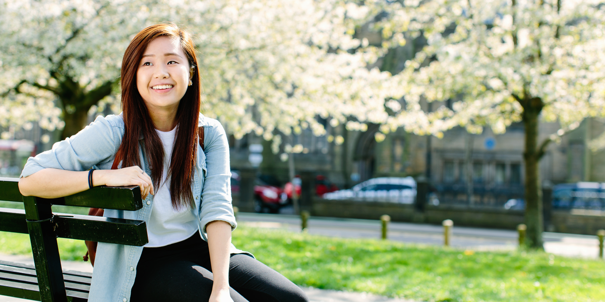 A student smiles sitting on a bench near cherry blossom trees.