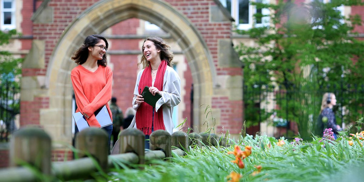 Two students chatting and laughing on campus. The Clothworkers Court is in the background and there are lots of flowers and greenery.