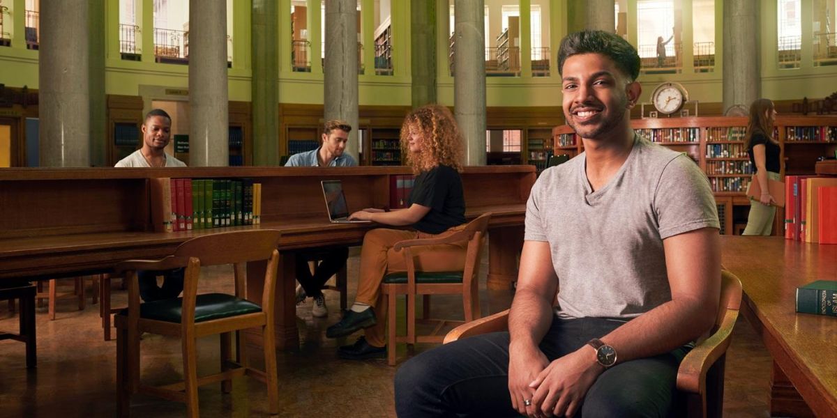 Seeveral students in tbhe Brotherton library working at desks with a student in the foreground smiling at the camera. The library interior is traditional in appearance with wooden furniture, bookshelves and stone pillars.