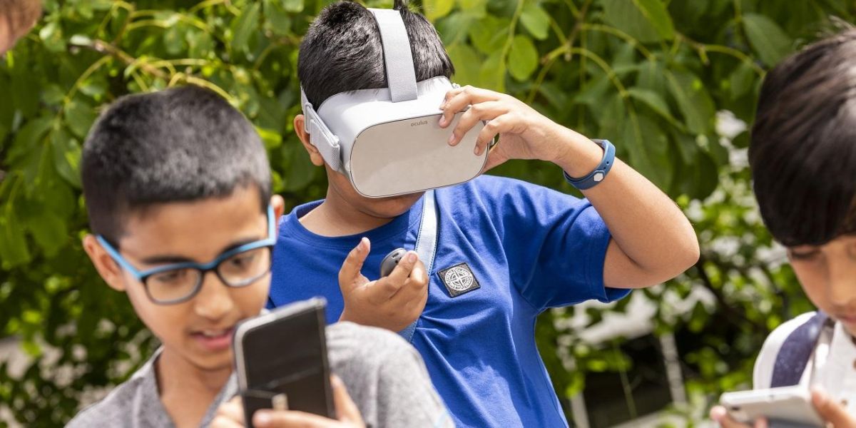 Three children in a park, two are looking at smartphones, one is wearing a VR headset