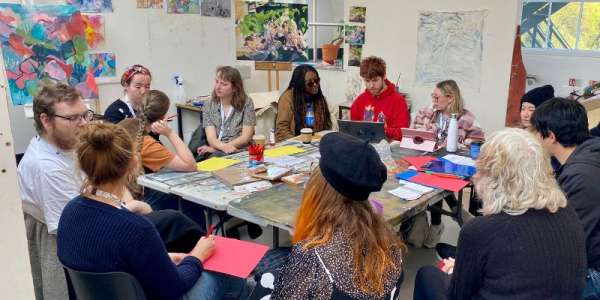 A group of artists are pictured gathered around a table, talking to each other. The walls are covered in colourful art pieces.