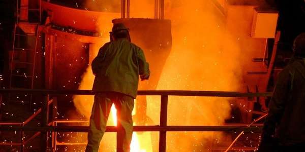 The picture shows a steel workier looking at a giant furnace as hot, molten metal is poured into it. The heat is so intense, an orange g glow is given off.