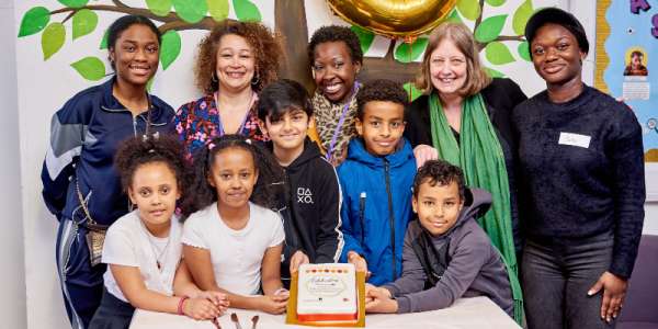 A group of adults and young people looking and smiling at the camera with a cake decorated with the words Celebrating IntoUniversity Leeds South belated 5th anniversary