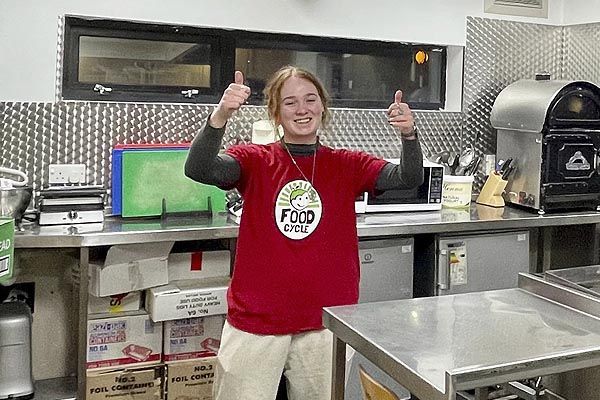 Anouska in a t-shirt that reads "Food Cycle", stood smiling in a large kitchen area.